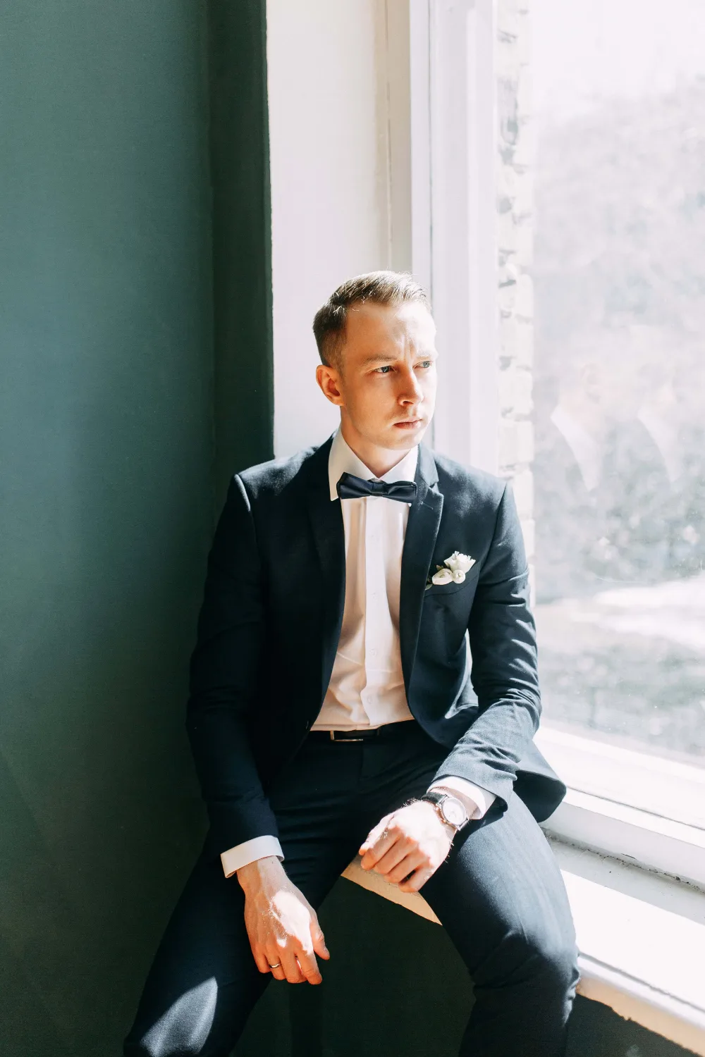 Groom wearing a navy blue wedding tuxedo by a window